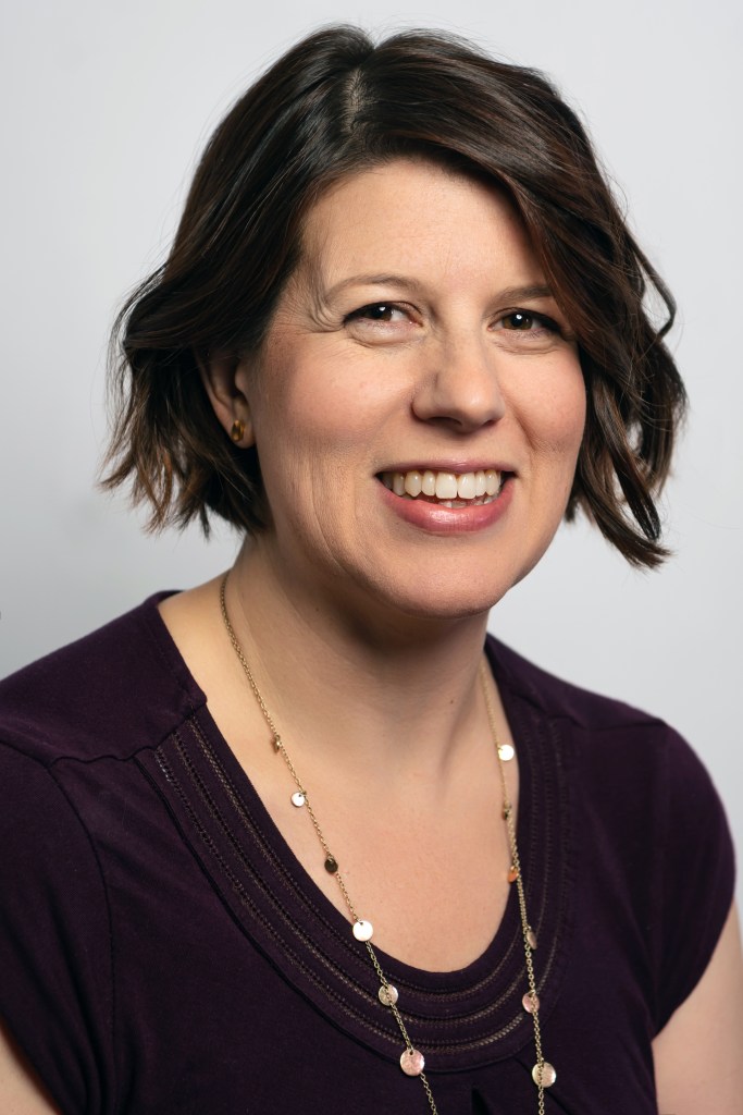 headshot of Carolyn. She is a white woman. Her hair was chin-length, brown, and curly on the day the photo was taken. She is wearing a maroon shirt and a long gold necklace.
